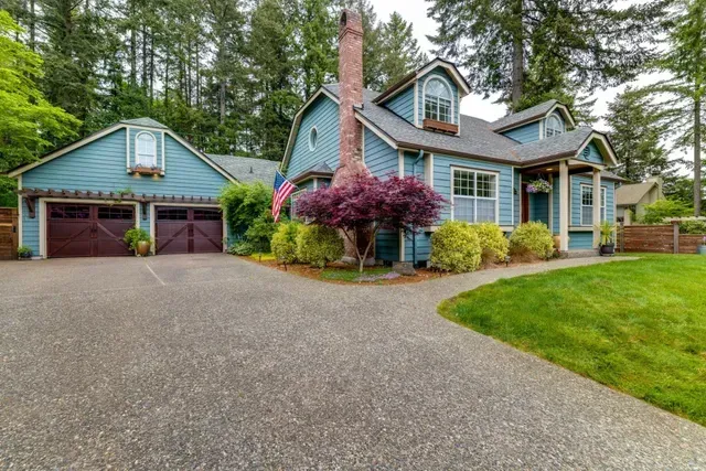 Blue house with garage, stone driveway, and lush green lawn surrounded by tall trees