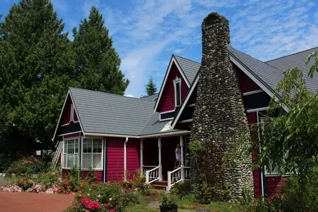 Red house with gray roof and stone chimney surrounded by trees and garden greenery.