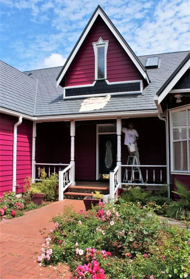 Bright pink house with a front porch, flower garden, and person standing in the doorway