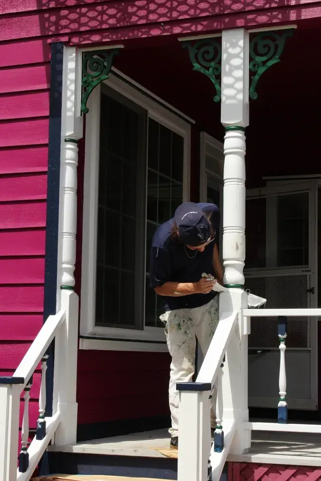Person standing on a porch, head down, working on a white railing beside a magenta house.