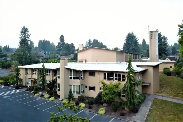 Low-rise beige office building with parking lot, trees, and a tall chimney-like tower