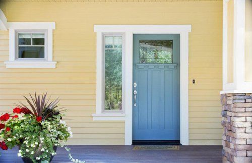 Blue front door with sidelight and small window, yellow siding, white trim, and a flower pot.