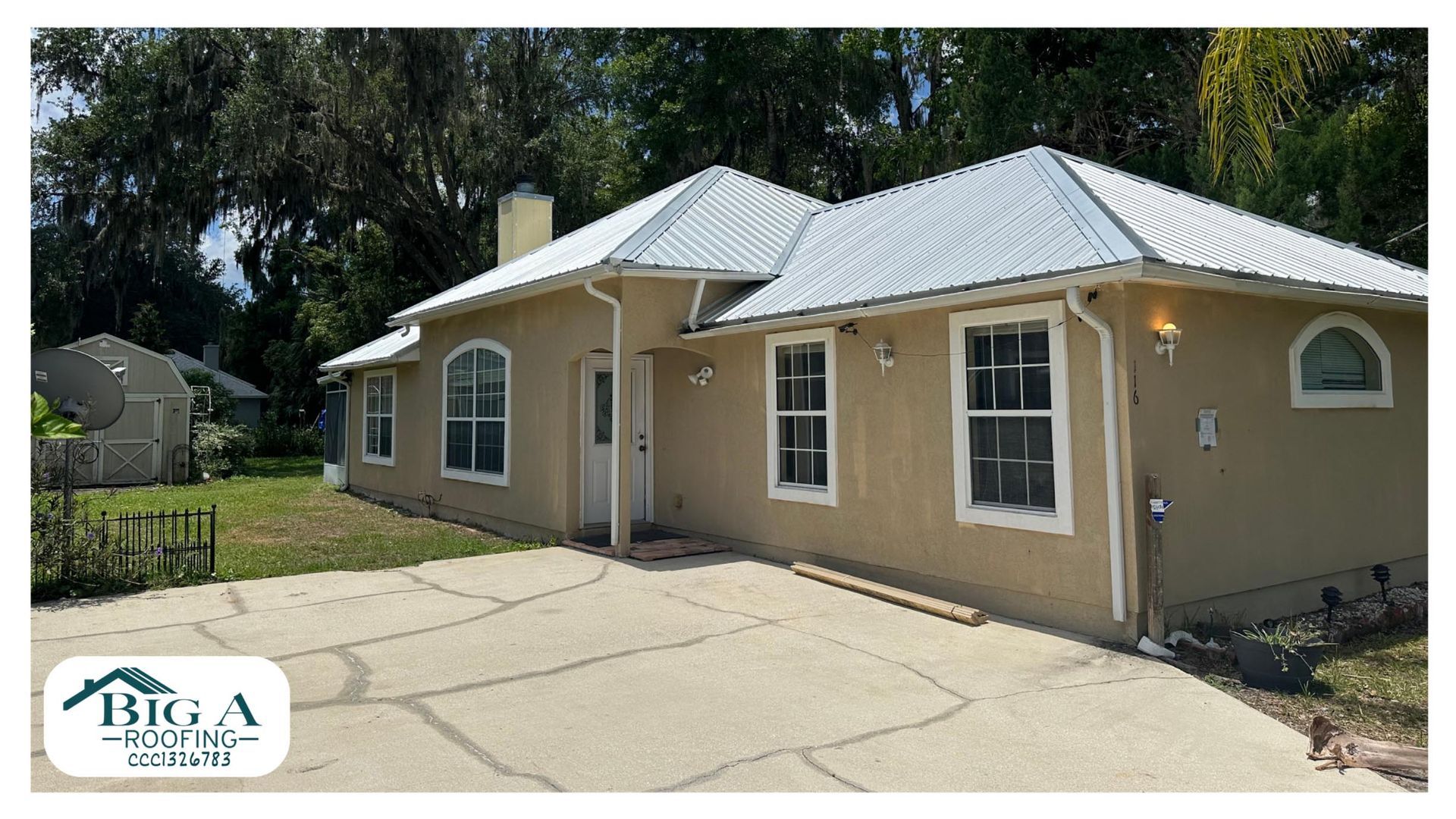 A tan, single-story house with a white metal roof and a cracked concrete driveway under a clear blue sky.