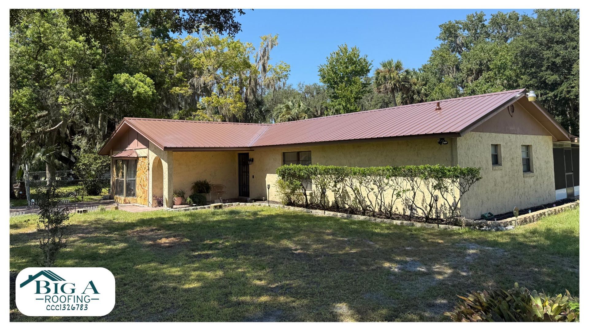 Single-story yellow house with a brown metal roof, surrounded by trees and a green lawn under a clear blue sky.