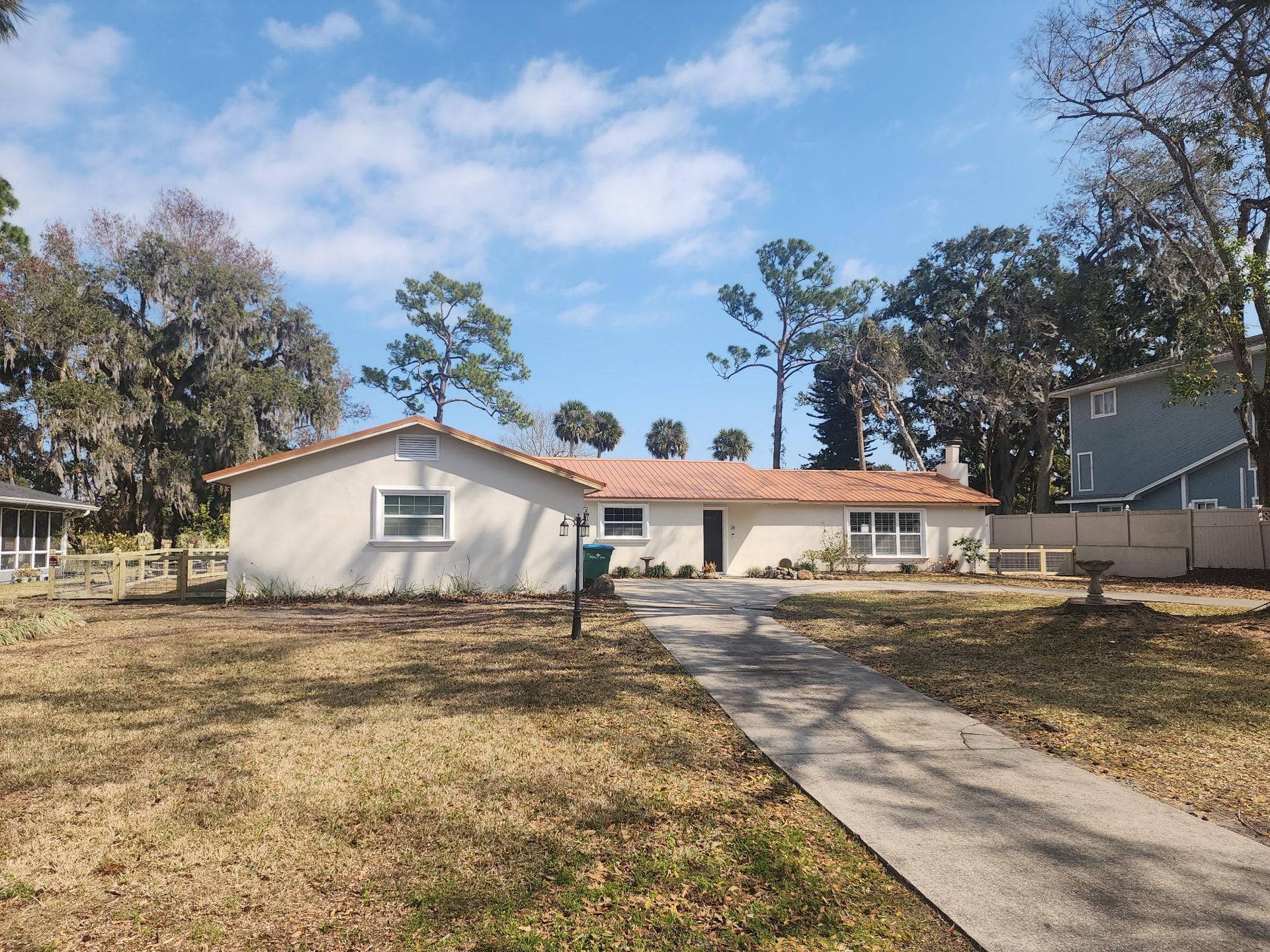 Single-story beige house with a terracotta tile roof, front sidewalk, and large trees under a blue sky.