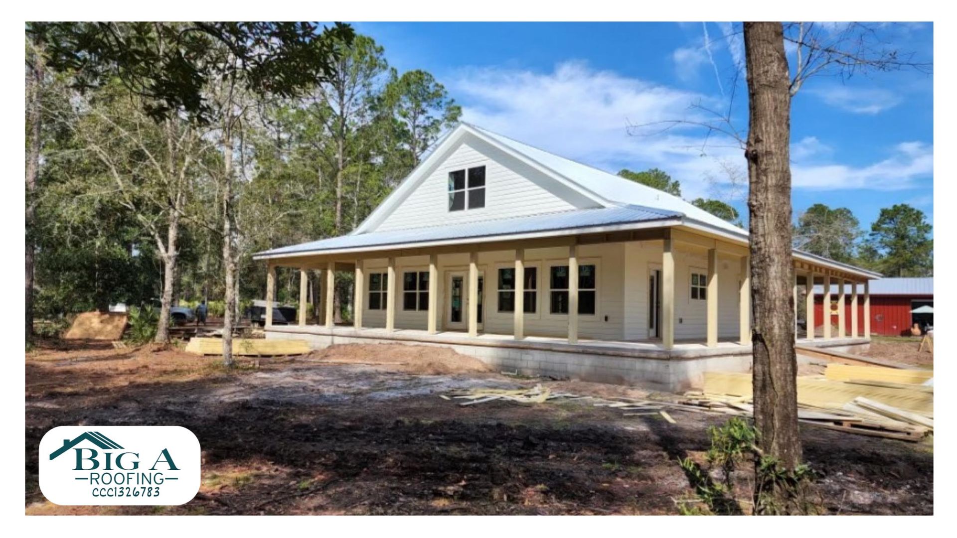 A modern white farmhouse with a large wraparound porch and a metal roof under construction in a wooded setting.