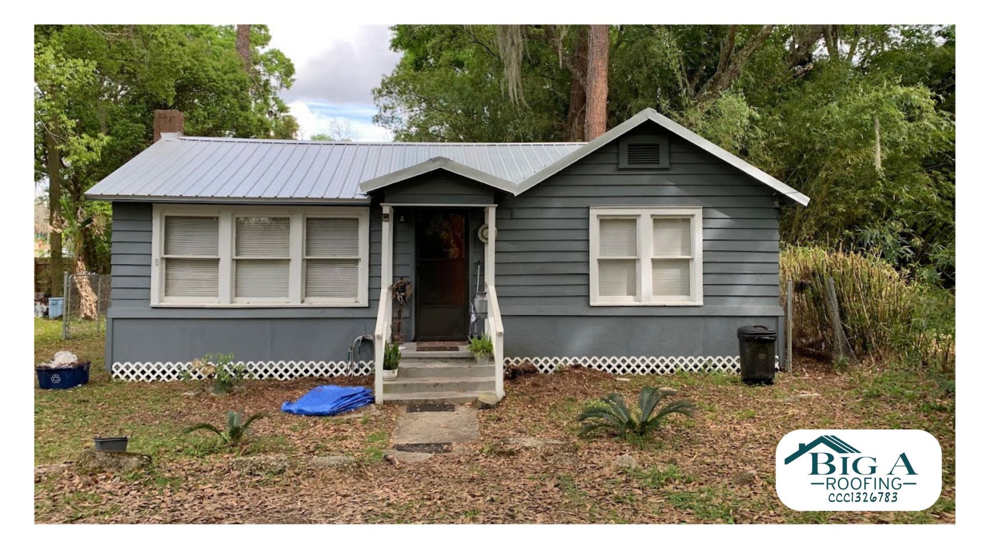A gray, single-story house with a light metal roof, white trim, and a covered front porch, surrounded by overgrown yard.