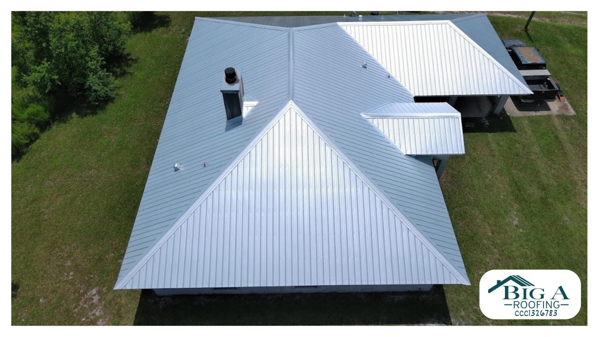 A top-down aerial view of a gray metal roof with a chimney and a small company logo in the corner.