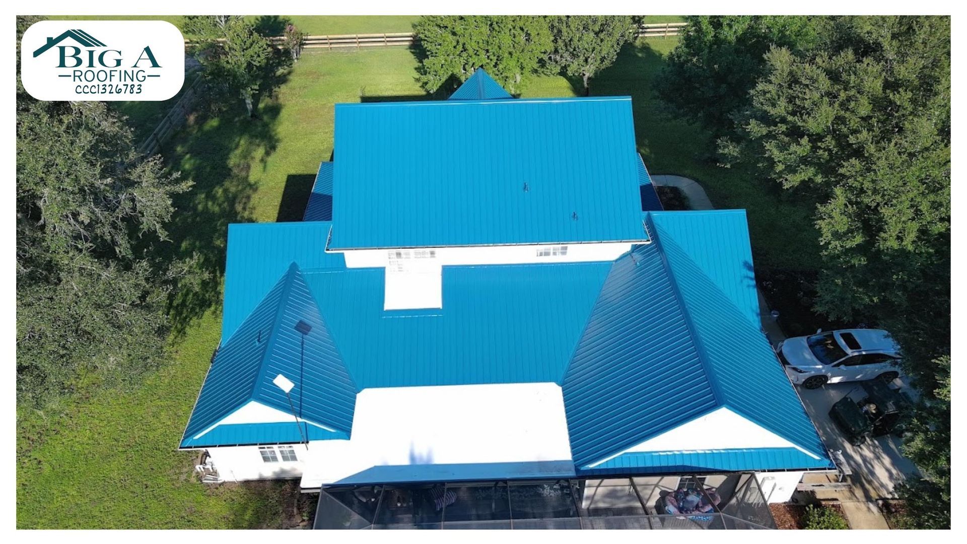 Aerial view of a house with a vibrant blue metal roof, surrounded by green trees, with a company logo in the corner.