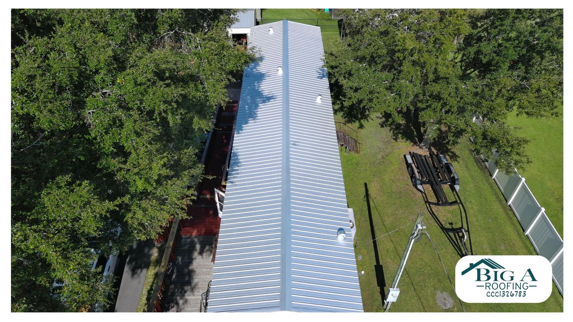 An aerial view of a long mobile home roof with a light-colored, patterned coating, next to trees and a utility trailer.
