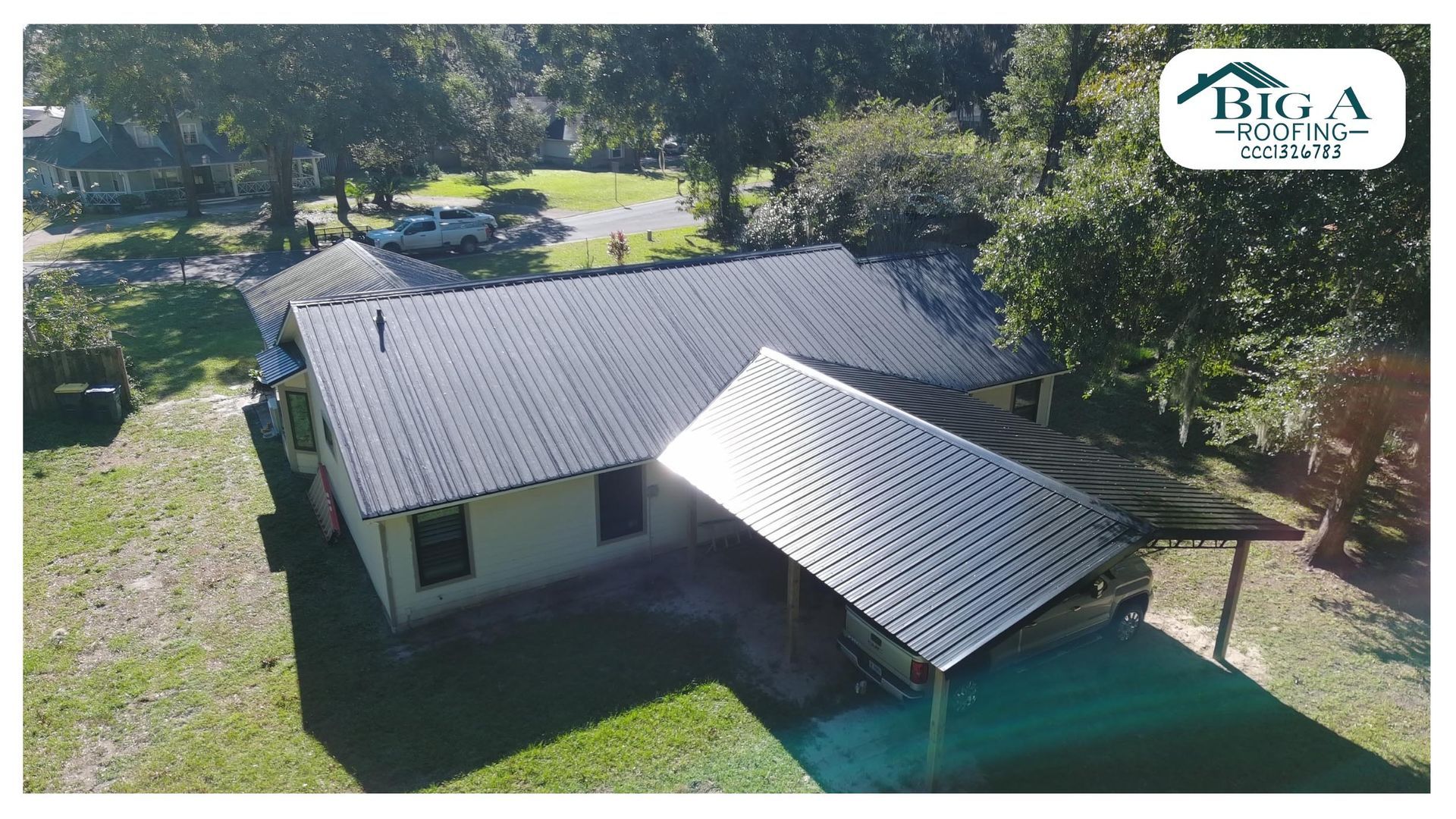 Aerial view of a light-colored house with a dark metal roof and a carport, surrounded by trees and a lawn.
