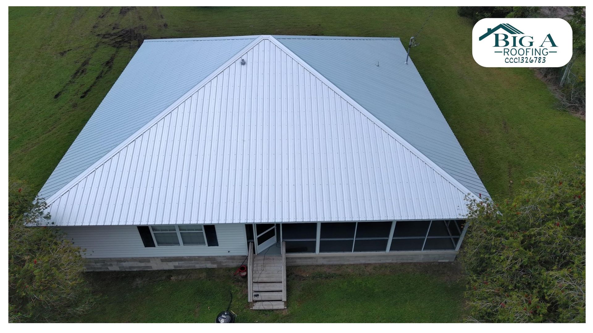 Aerial view of a white single-story house with a grey metal roof and a screen-enclosed patio, surrounded by a green lawn.