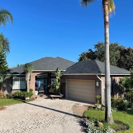 A brown single-story house with a gray roof and a tan garage door sits behind a gravel driveway under a clear blue sky.