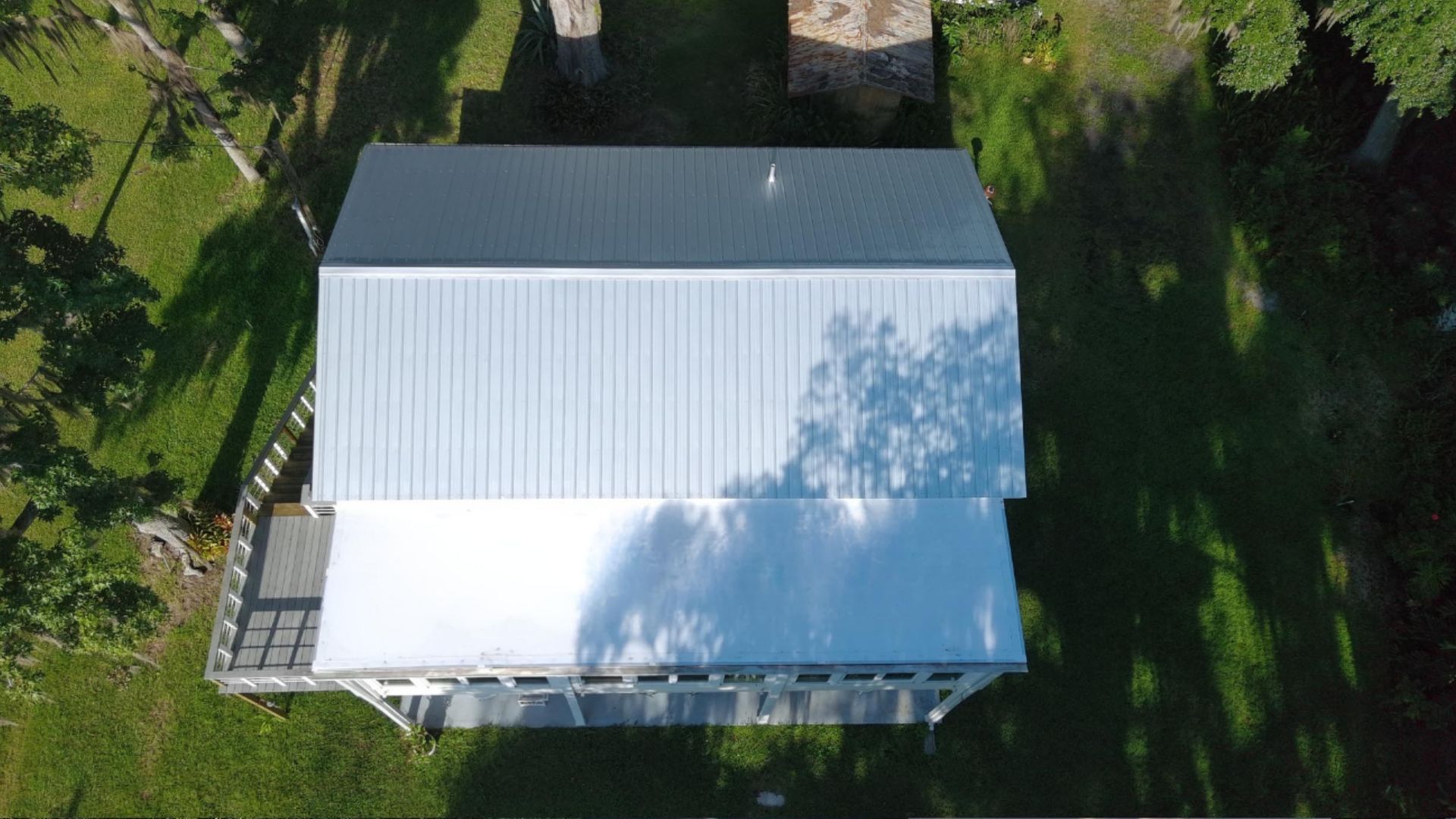 An aerial view of a rectangular house with a light-colored metal roof, surrounded by a green, tree-lined lawn.