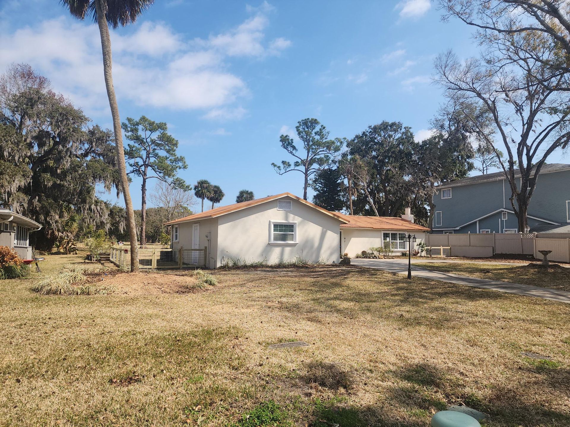 A light-colored, single-story house with a tan tile roof sits on a large, dry lawn under a blue sky with scattered clouds.