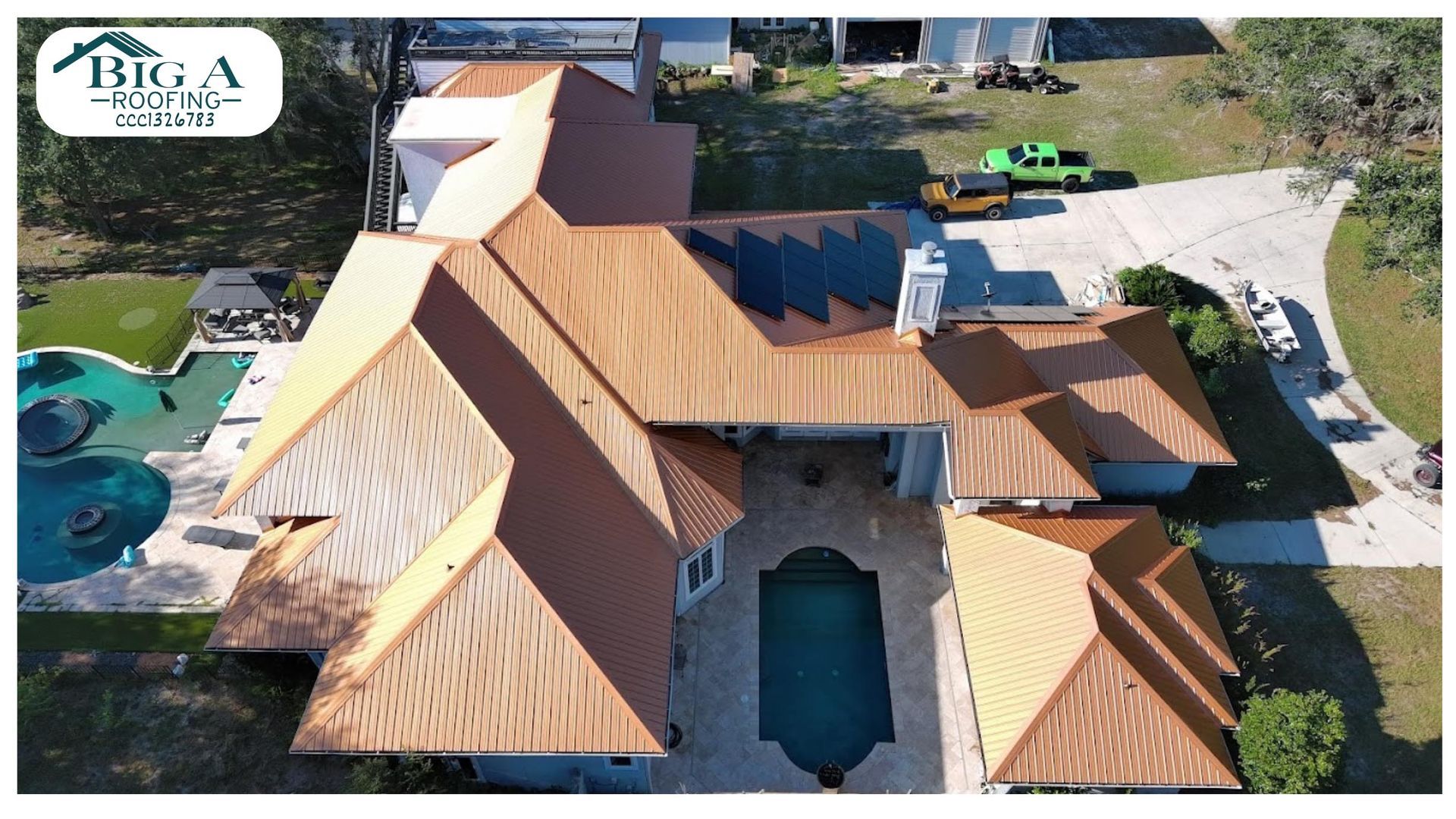 Aerial view of a large residential house with a new tan metal roof, featuring a pool and driveway.