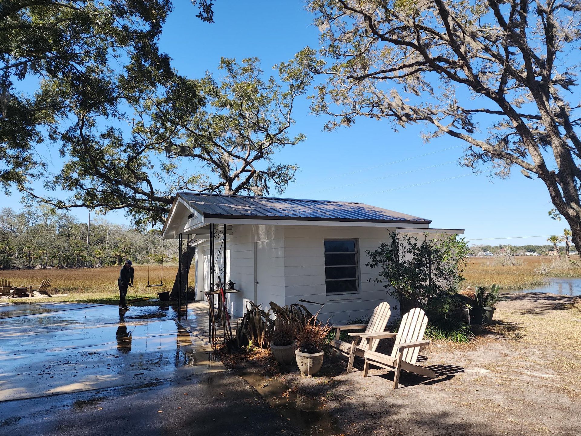 A white cottage stands by a marshy waterfront on a sunny day, with two wooden chairs outside and a person nearby.