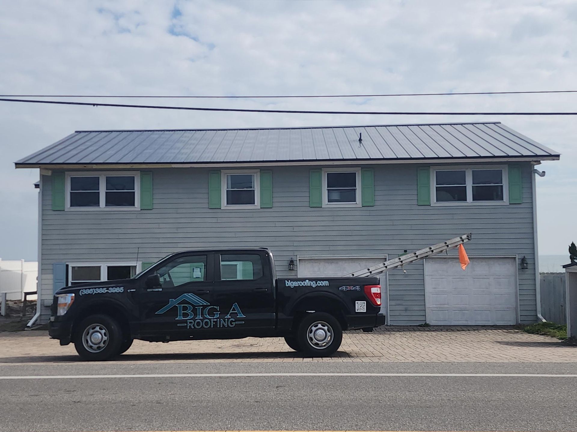 A black pickup truck with a ladder parked in front of a gray two-story building with a metal roof under a cloudy sky.