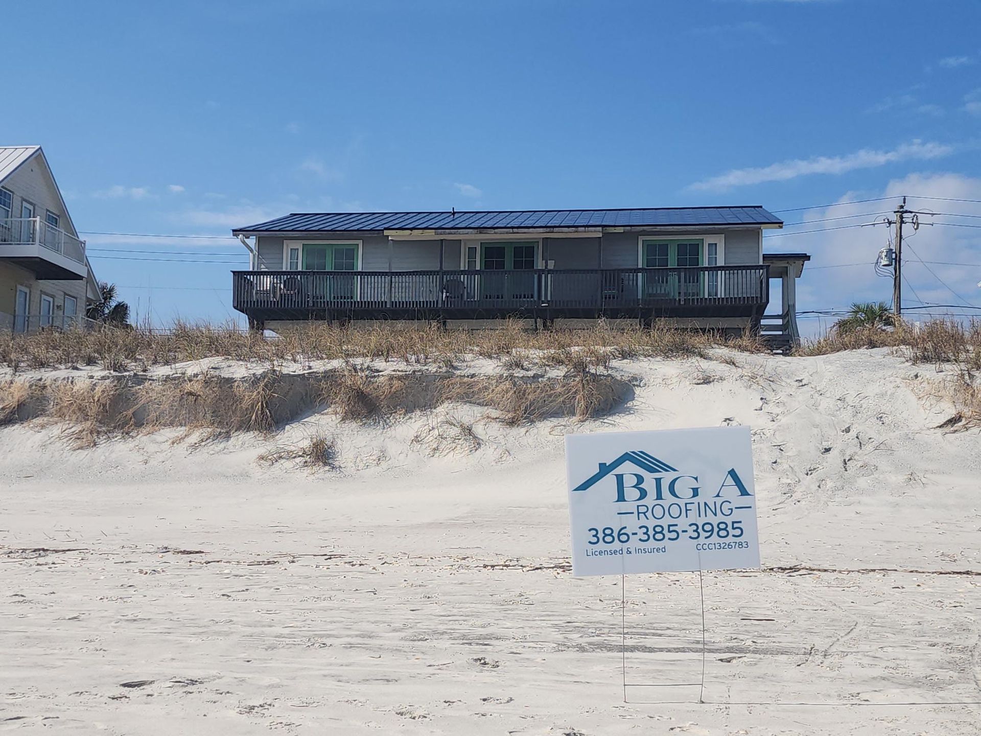 A beachfront house with a metal roof and deck sits above sandy dunes, with a Big A Roofing sign in the foreground.