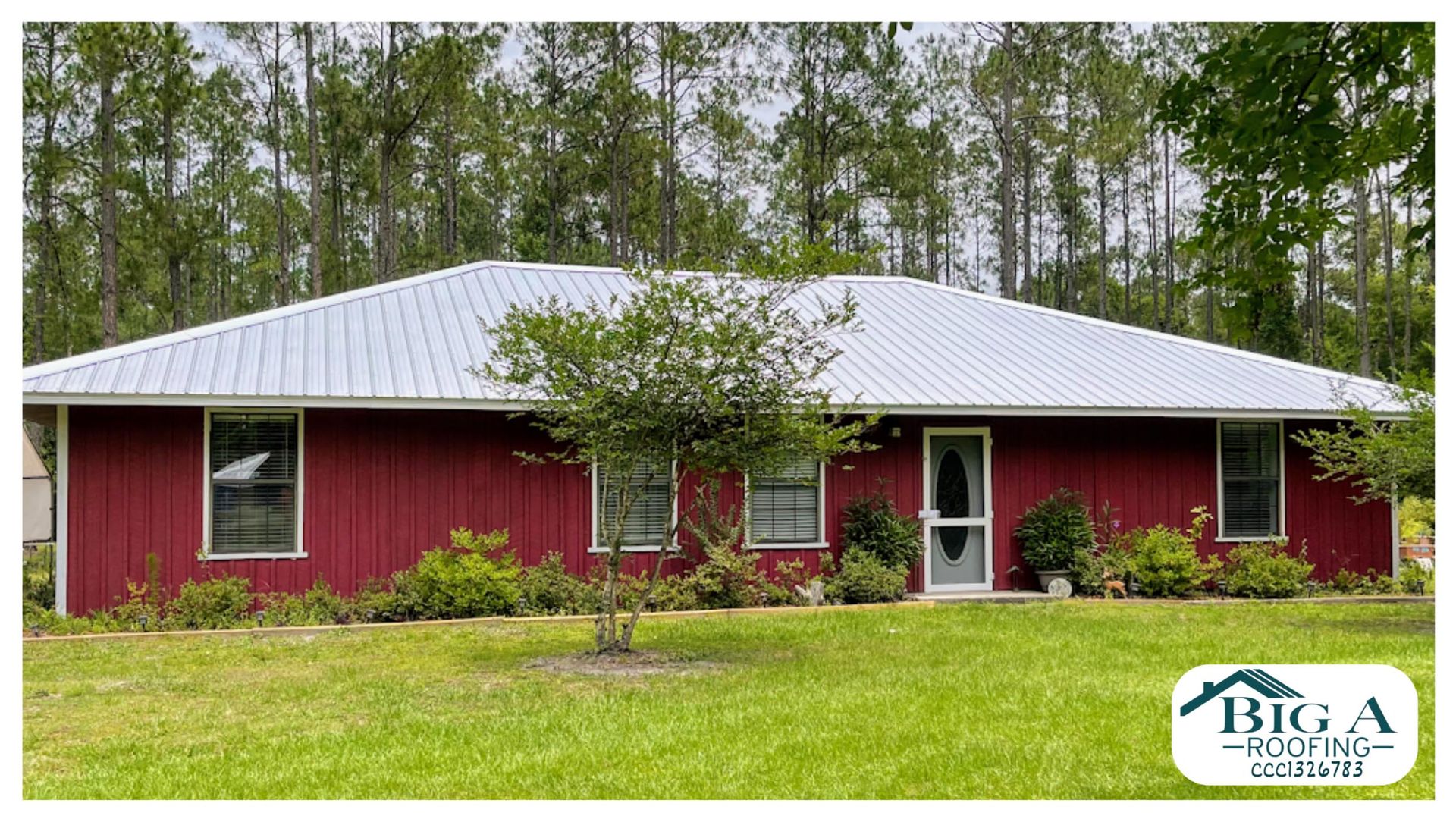 A red house with a light-colored metal roof, surrounded by green trees and lawn, with a small business logo in the corner.