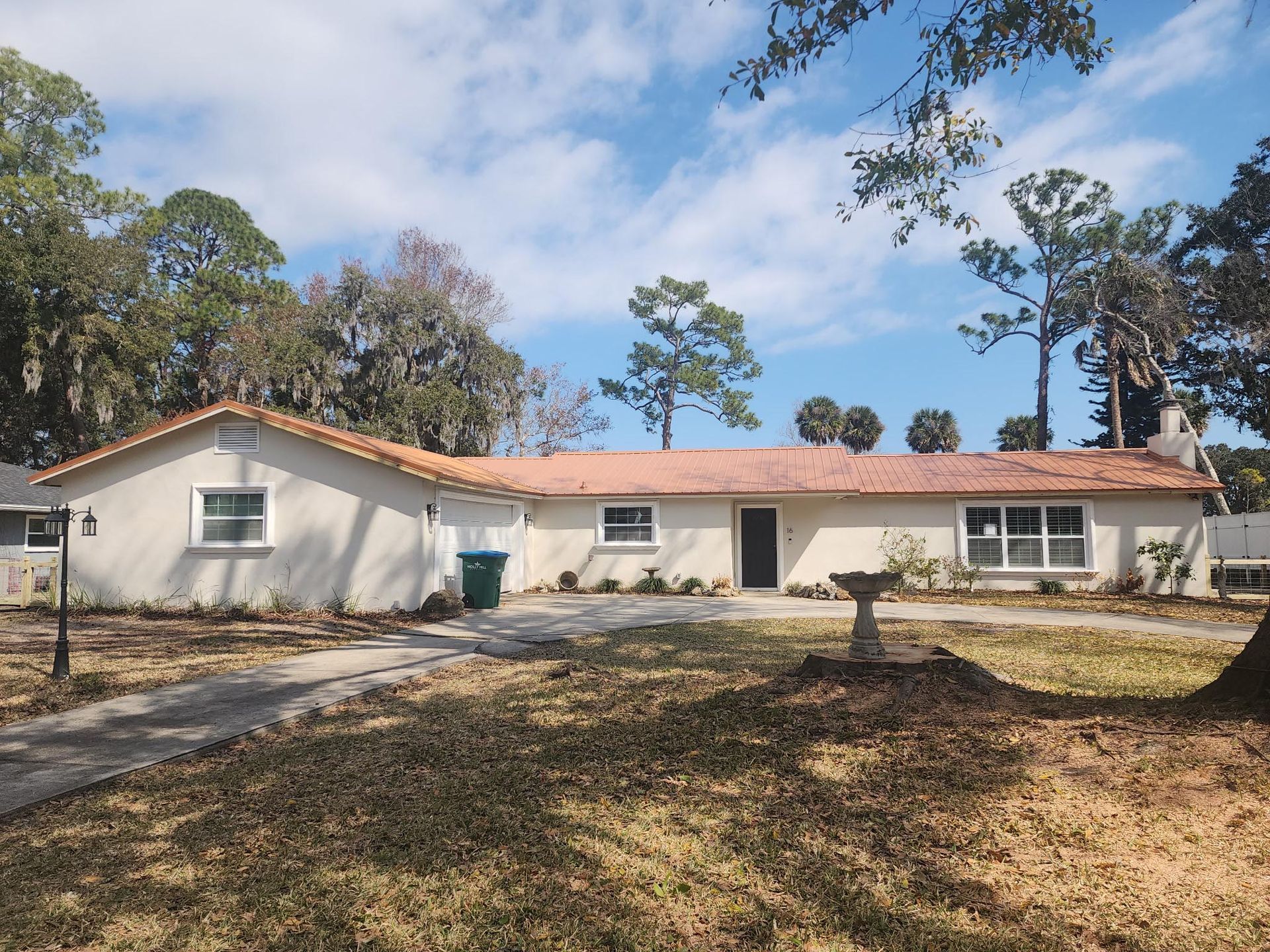 A light-colored, single-story ranch house with a reddish-orange tile roof under a partly cloudy sky with trees.