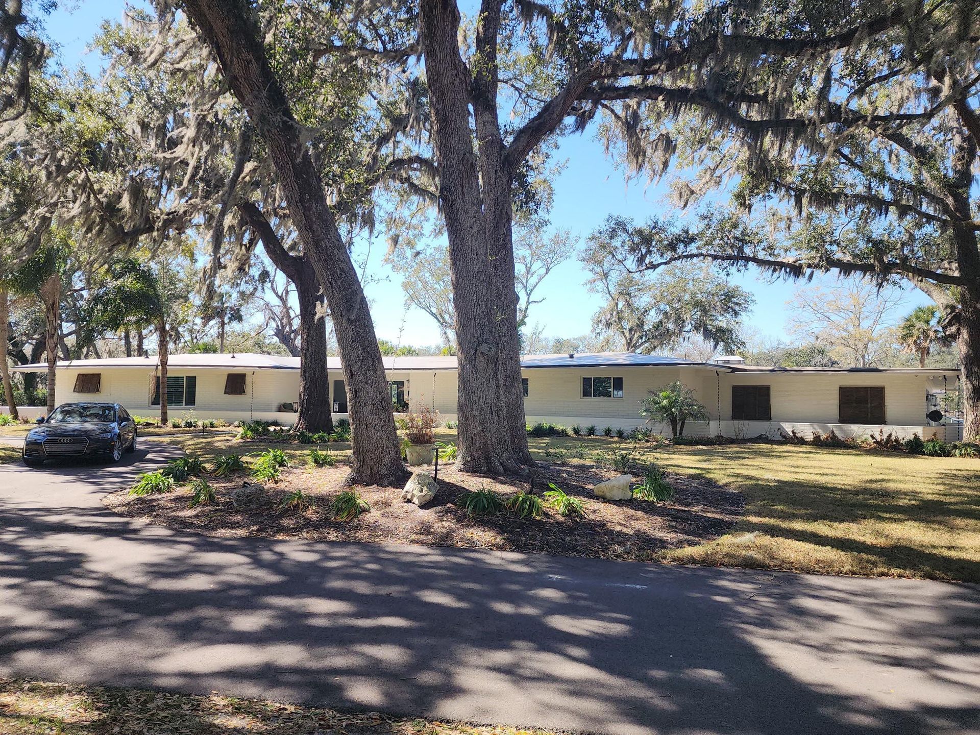 A long, single-story, beige ranch-style house nestled behind two large oak trees on a sunny, grassy property.