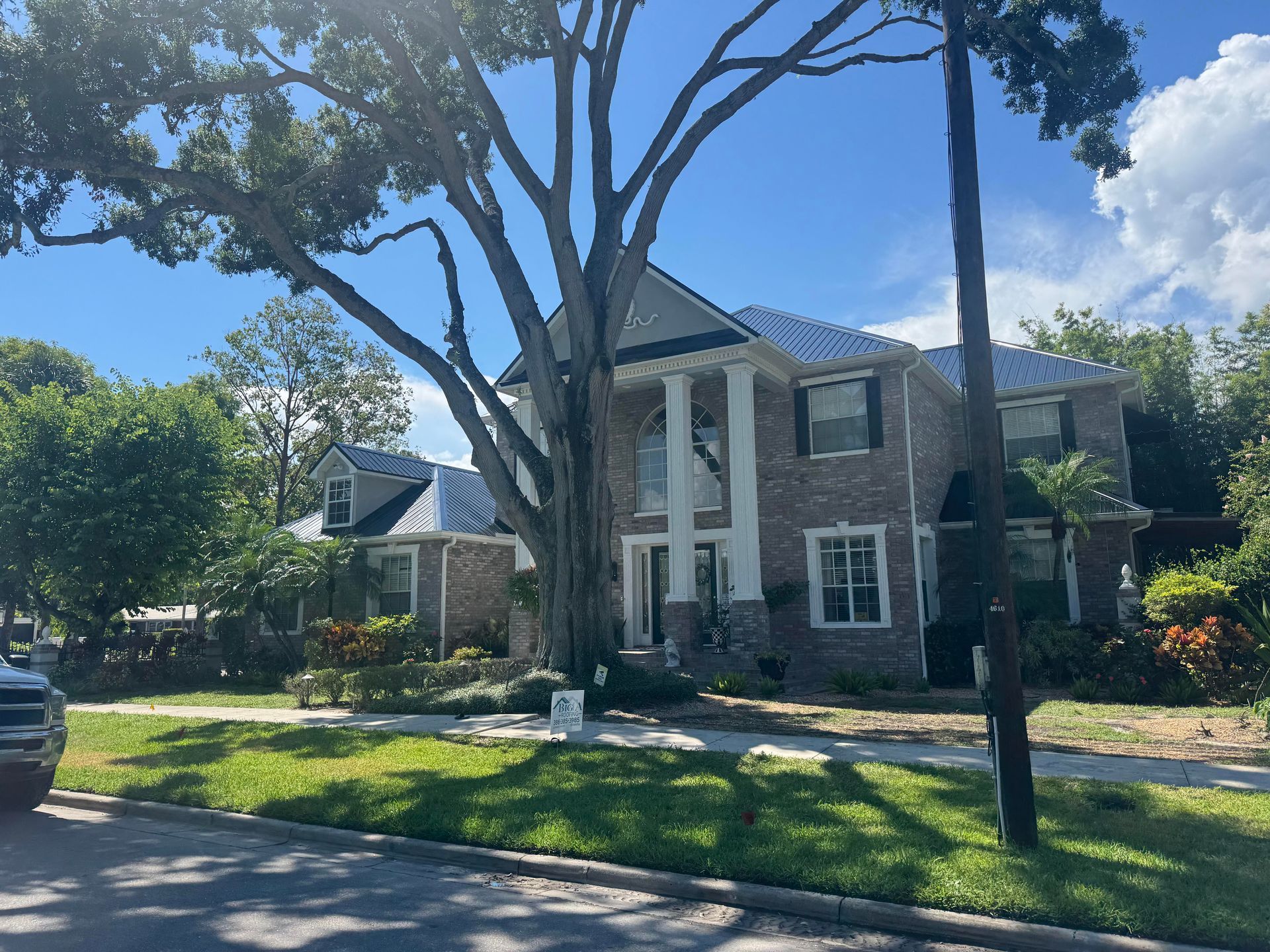 A two-story brick house with white columns and a metal roof, partially obscured by a large tree in a sunny suburban yard.