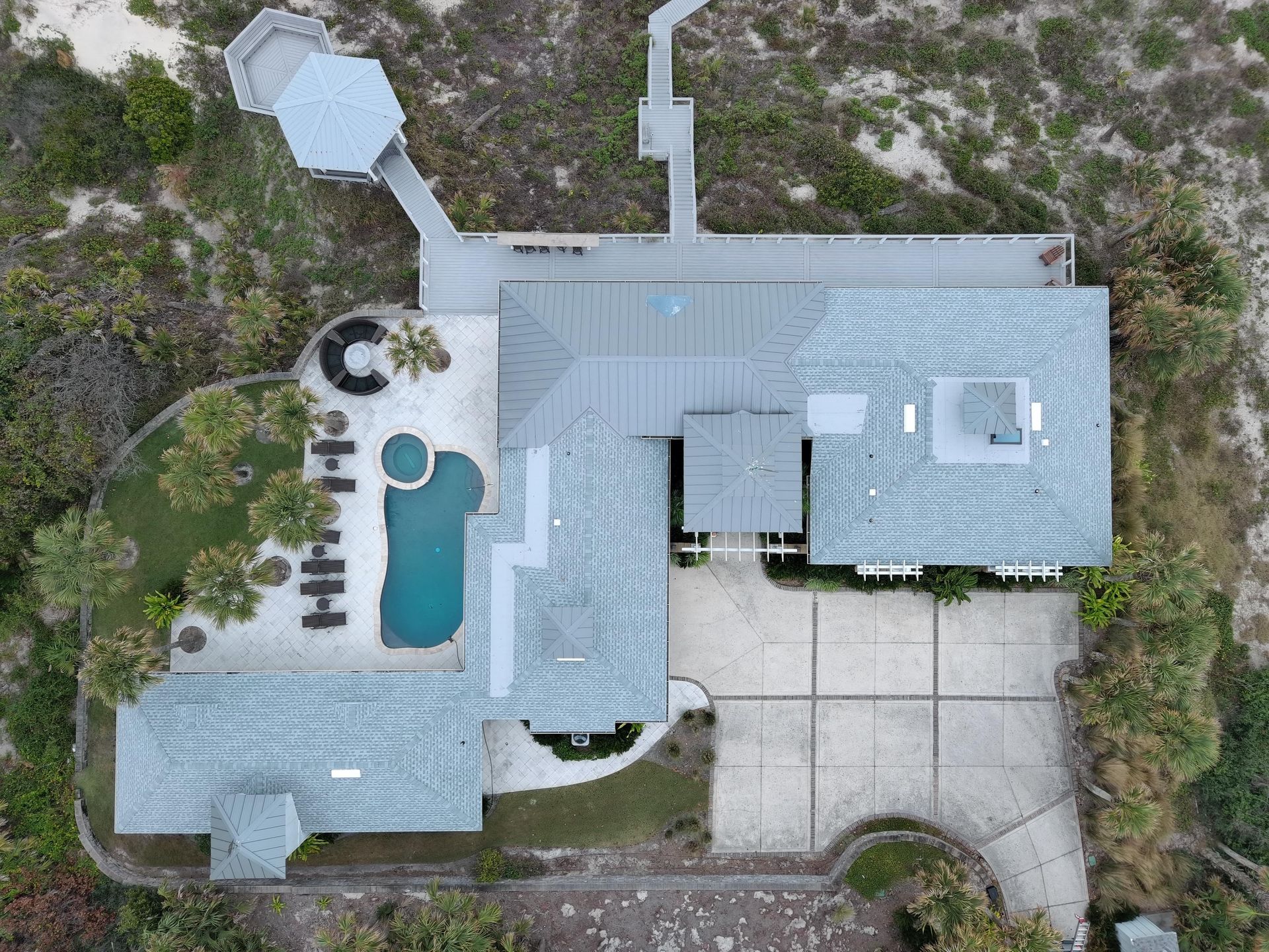 An aerial view of a coastal home with a pool, patio, and a boardwalk leading toward dunes.