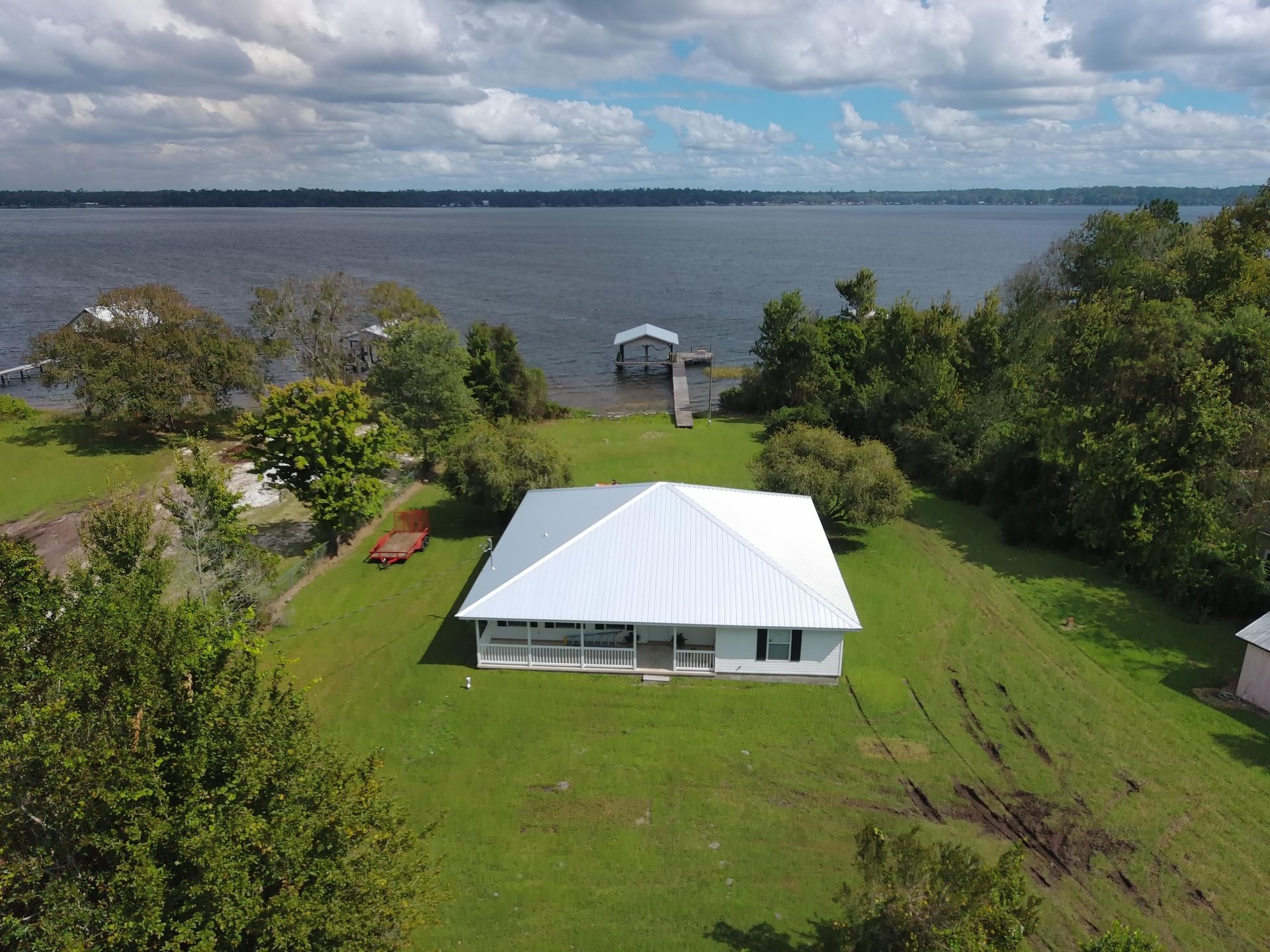 A white house with a metal roof sits on a green lawn by a large lake, featuring a private pier and a boat lift.