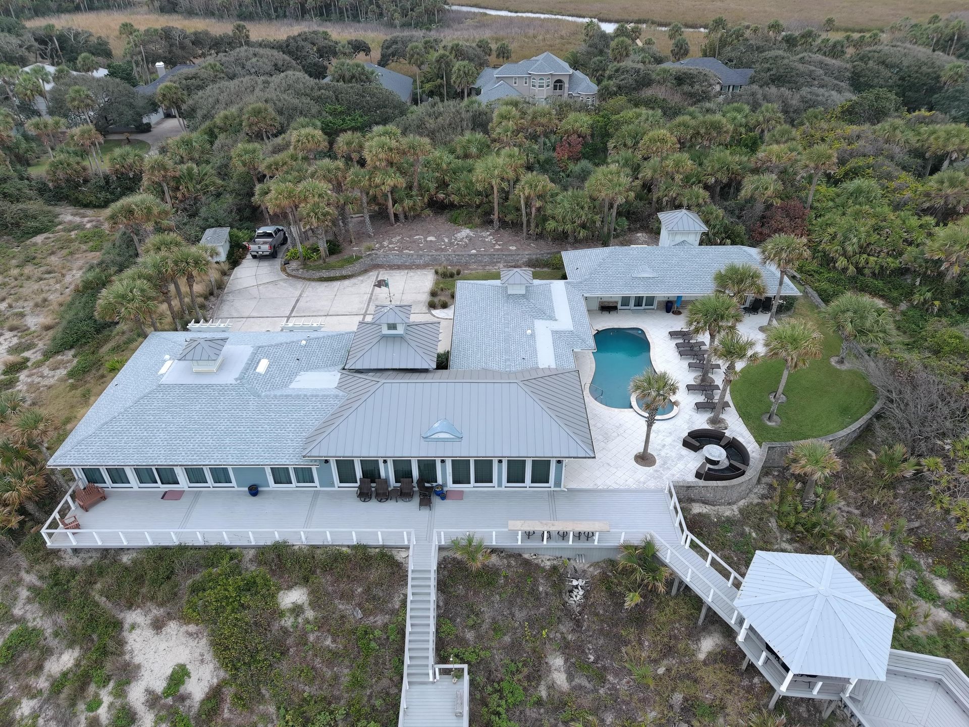 Aerial view of a coastal home with a grey metal roof, swimming pool, and deck, surrounded by lush vegetation.