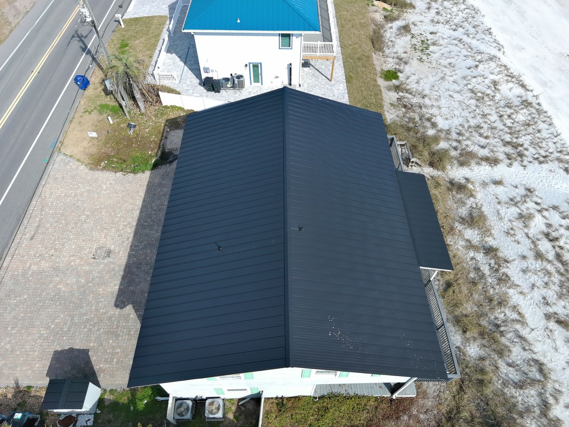 Aerial view of a rectangular, dark metal roof on a building, located near a road and a light-colored beach area.