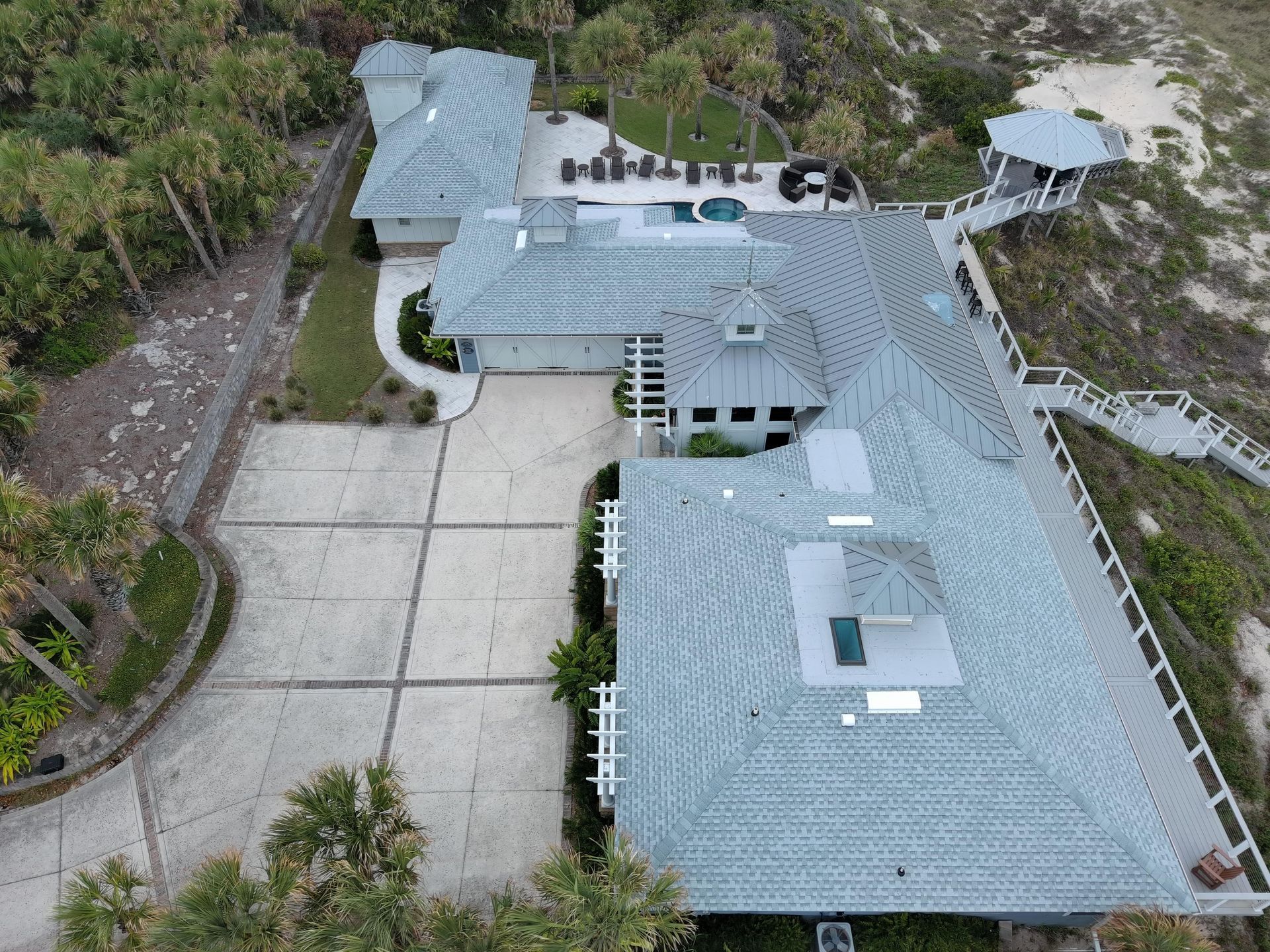 Aerial view of a coastal house with a light gray tiled roof, concrete driveway, swimming pool, and a walkway to the beach.