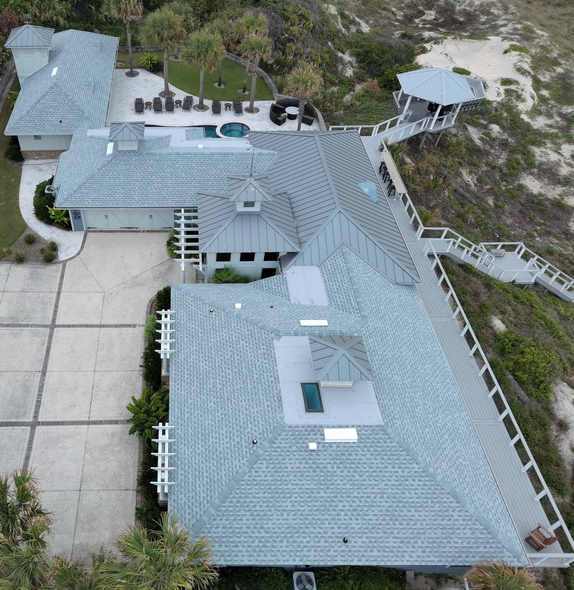 An aerial view of a large coastal home with light gray roofs, a swimming pool, and a walkway leading to a beach gazebo.