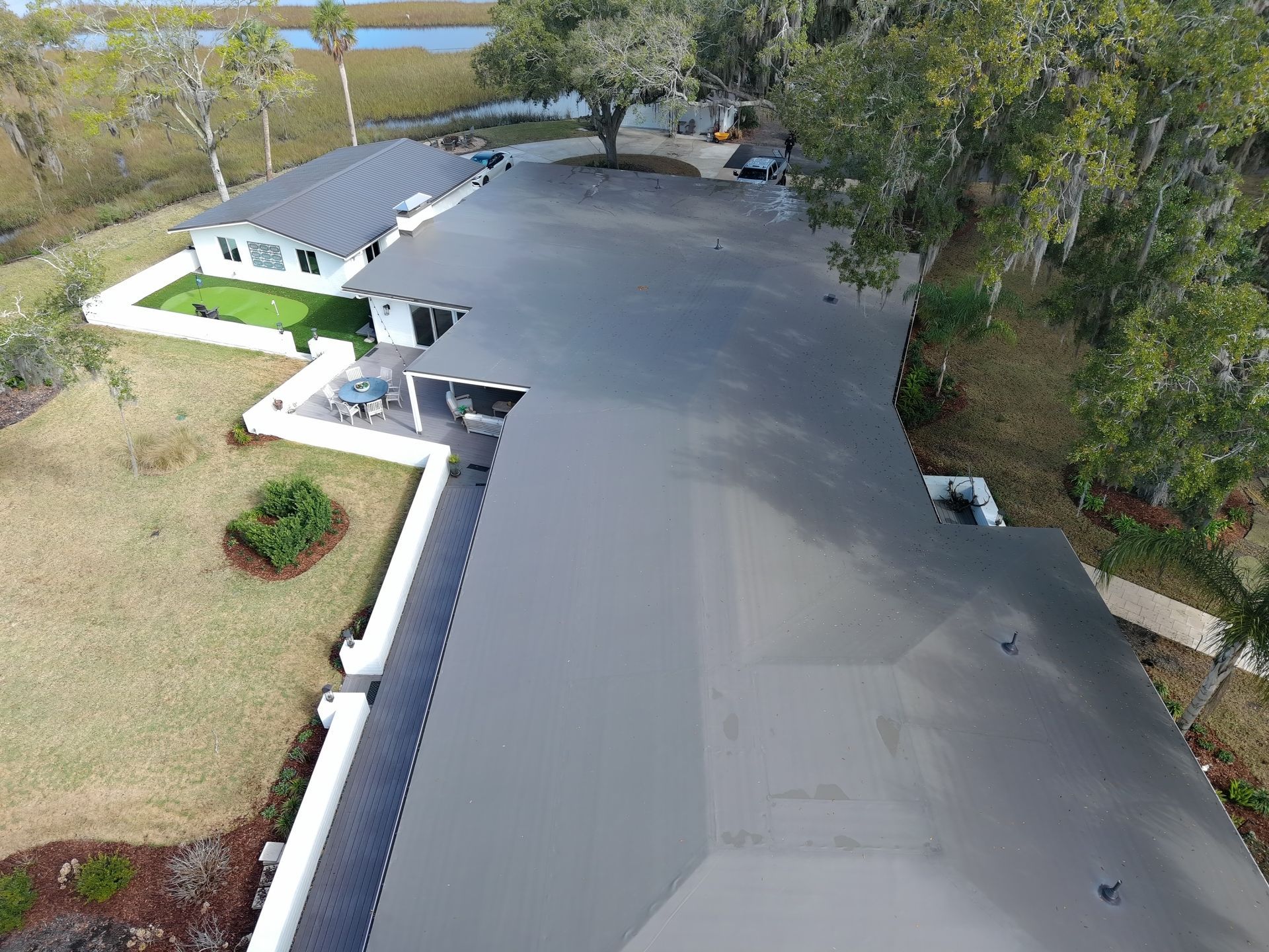 An aerial view of a single-story house with a flat, grey roof, surrounded by a yard, white fencing, and nearby trees.