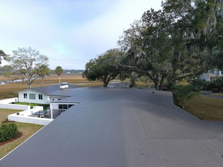 A view from above showing a flat gray roof extending over a house near a coastal marsh and large, mossy oak trees.