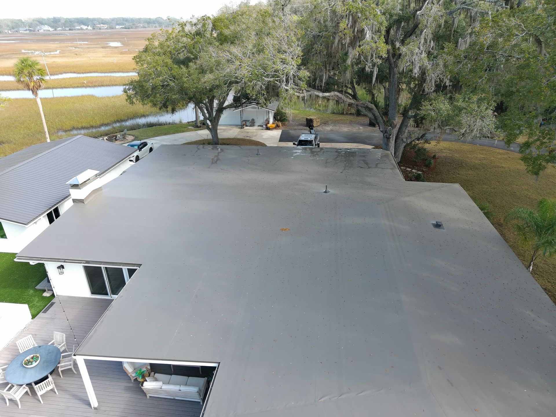 Aerial view of a flat grey roof on a white house, surrounded by trees and a marshland landscape.
