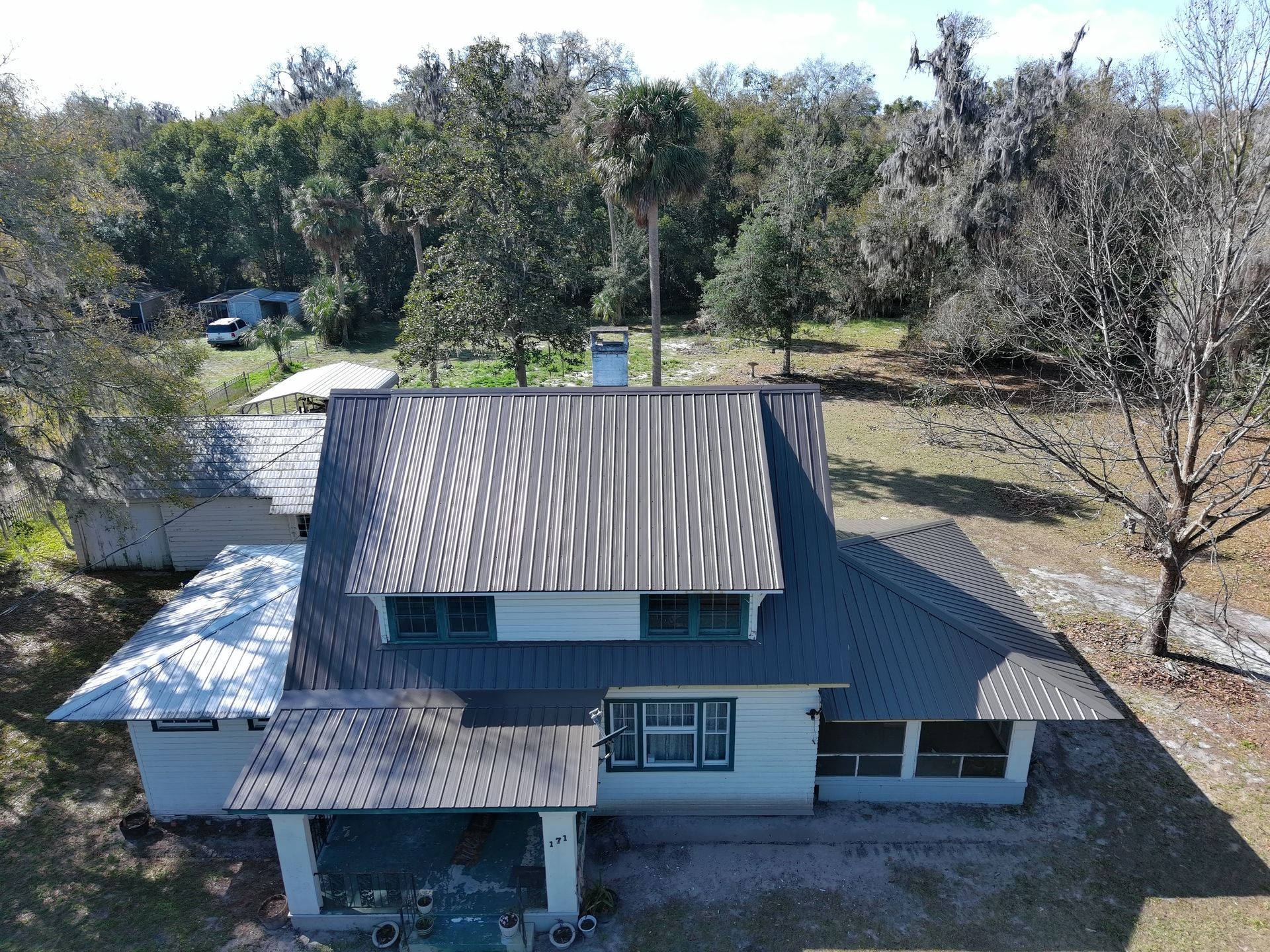 Aerial view of a white, two-story house with a brown metal roof surrounded by trees and a yard.
