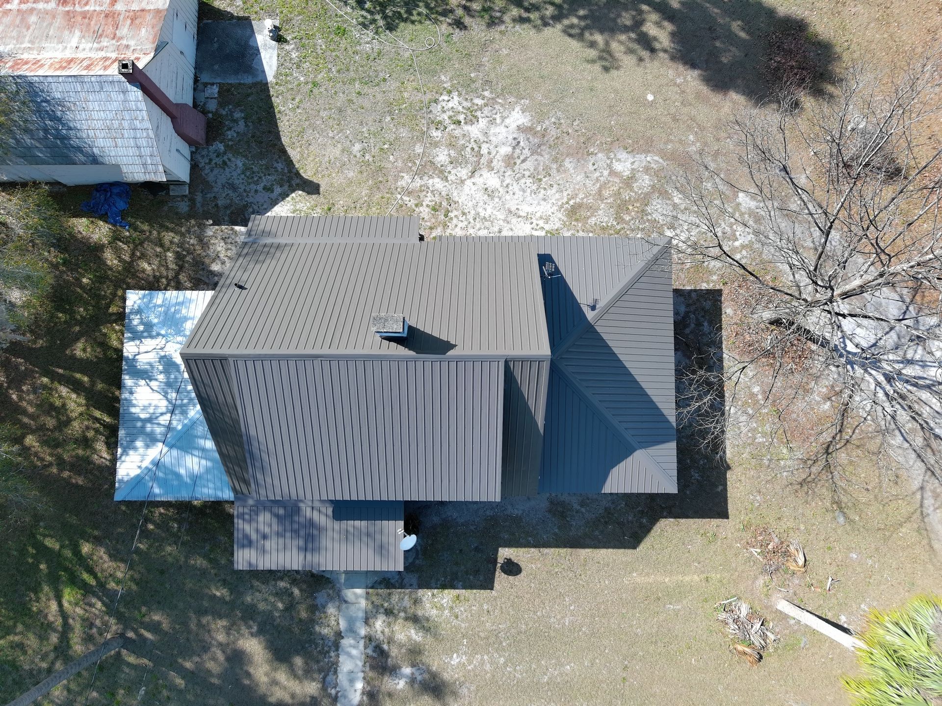 An aerial view of a small house with a grey metal roof, situated on a dry, grassy plot next to a white shed.