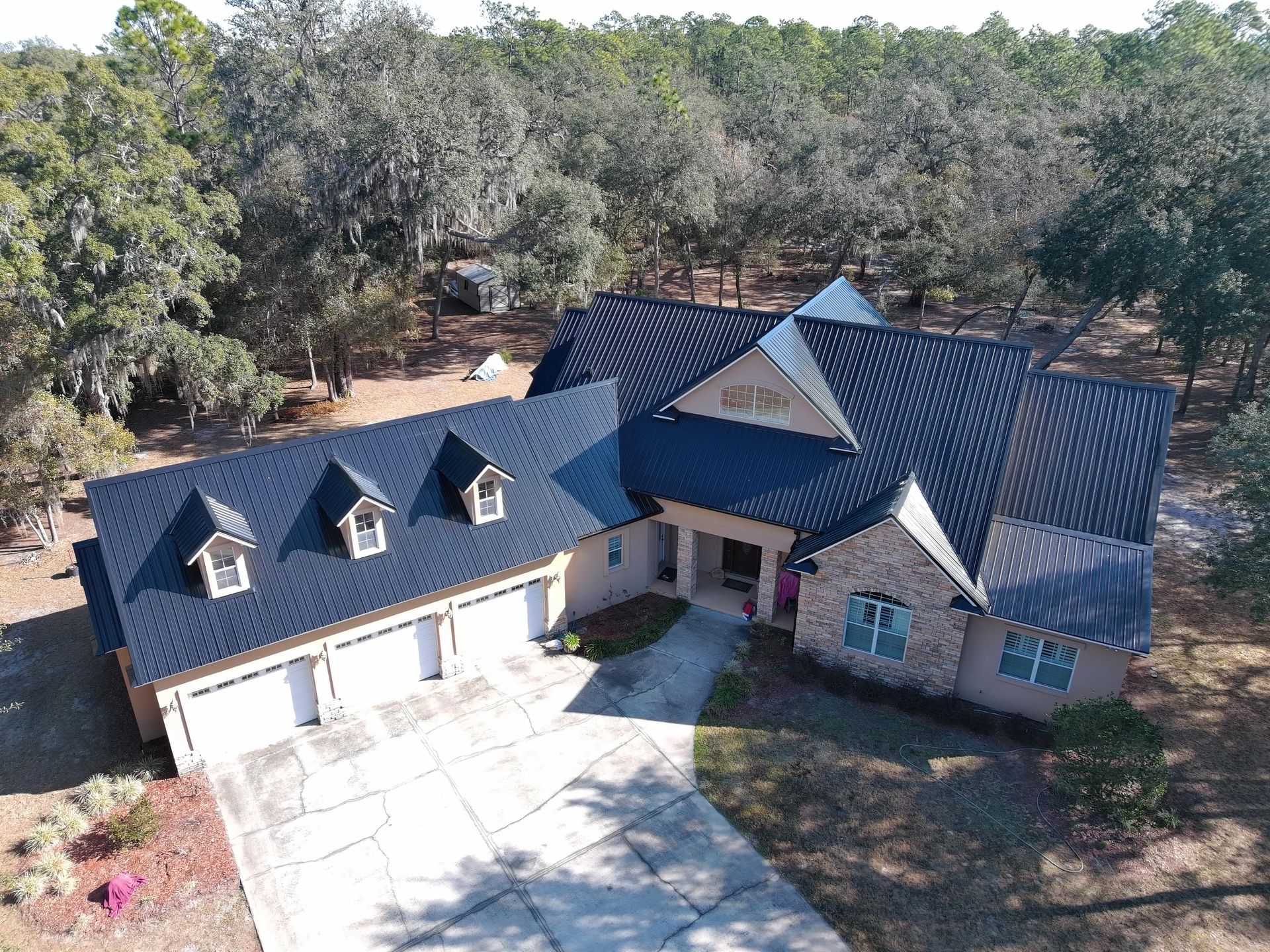 An aerial view of a single-story house with a dark metal roof, three garage bays, and a stone facade, set in a wooded area.