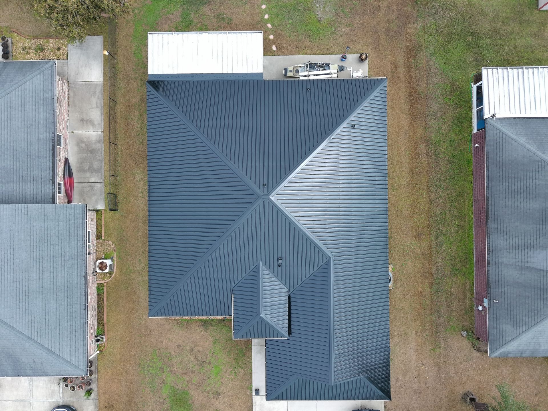 Aerial view of a residential dark metal hip roof with a small projecting front section, surrounded by grass and houses.