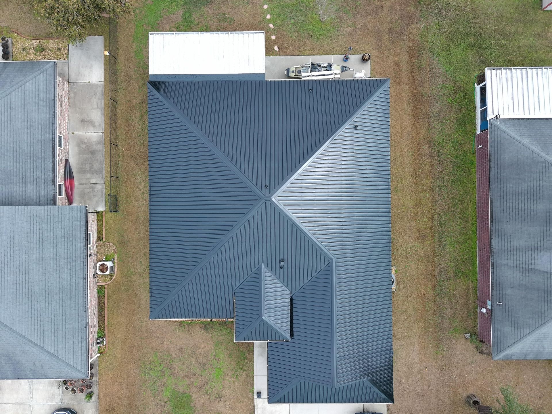 A bird's-eye view of a dark metal roof on a house, surrounded by neighboring residential roofs and patches of grass.