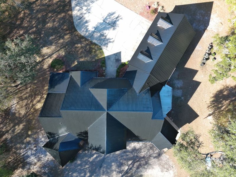 An aerial view of a dark metal-roofed house surrounded by trees, with a concrete driveway leading to the entrance.