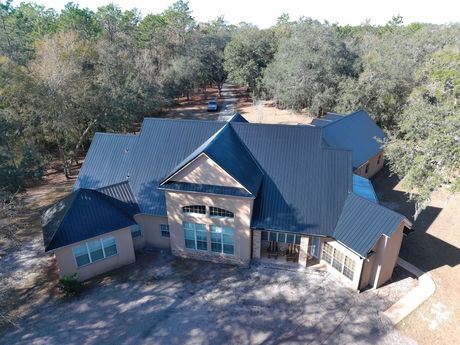 An aerial view of a tan-colored house with a dark metal roof, surrounded by trees on a wooded lot.