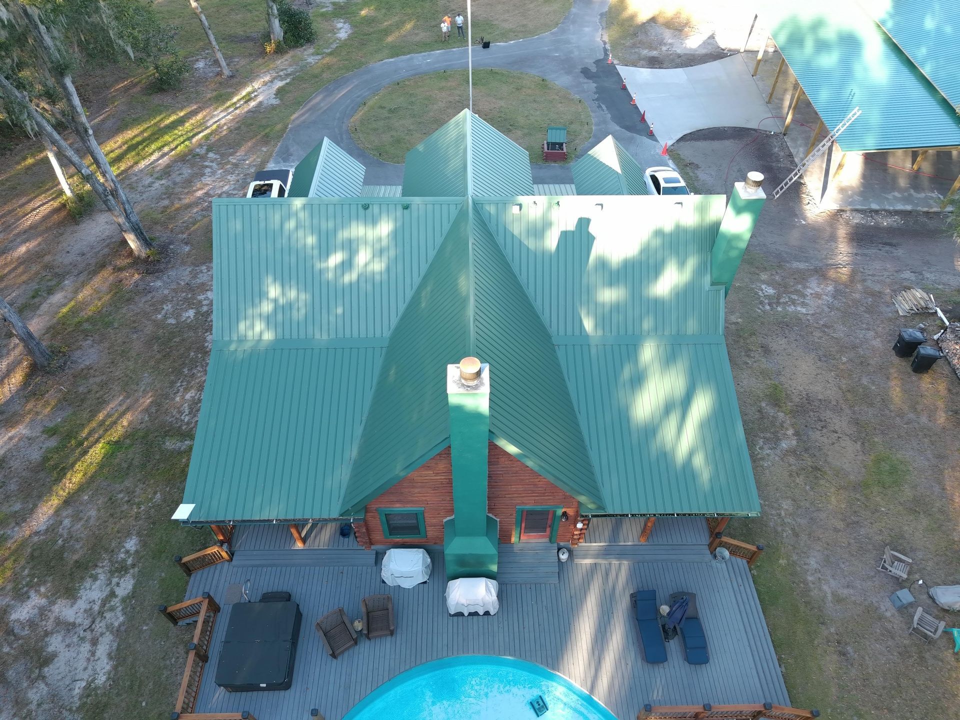 Aerial view of a cabin with a green metal roof, wrap-around deck, and backyard pool in a wooded area.