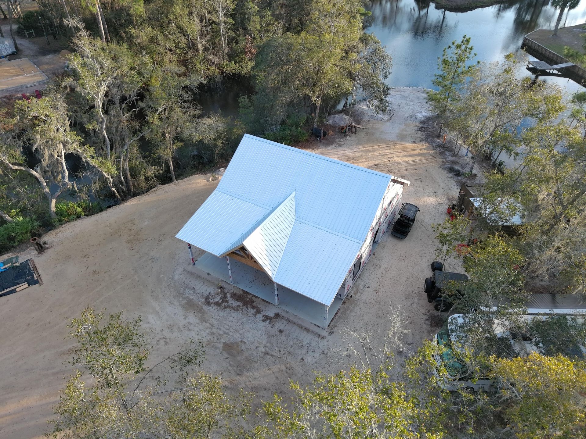 An aerial view of a metal-roofed building with an attached porch, situated on a sandy lot near trees and a body of water.