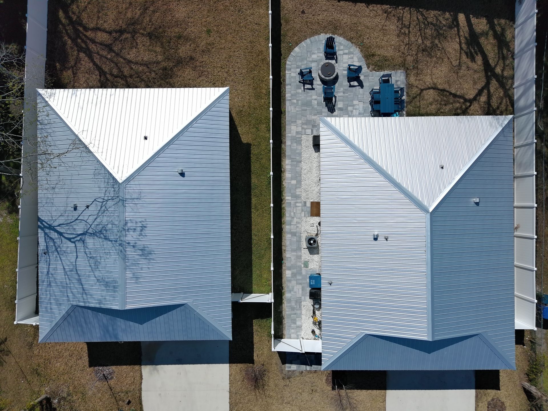 Aerial view of two homes with metal roofs, one featuring a paved patio area with outdoor furniture and a fire pit.