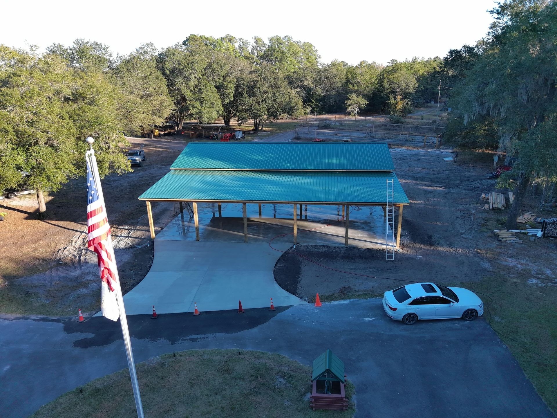An aerial view of a pavilion with a green metal roof, a concrete pad, a prominent American flag, and a white parked car.