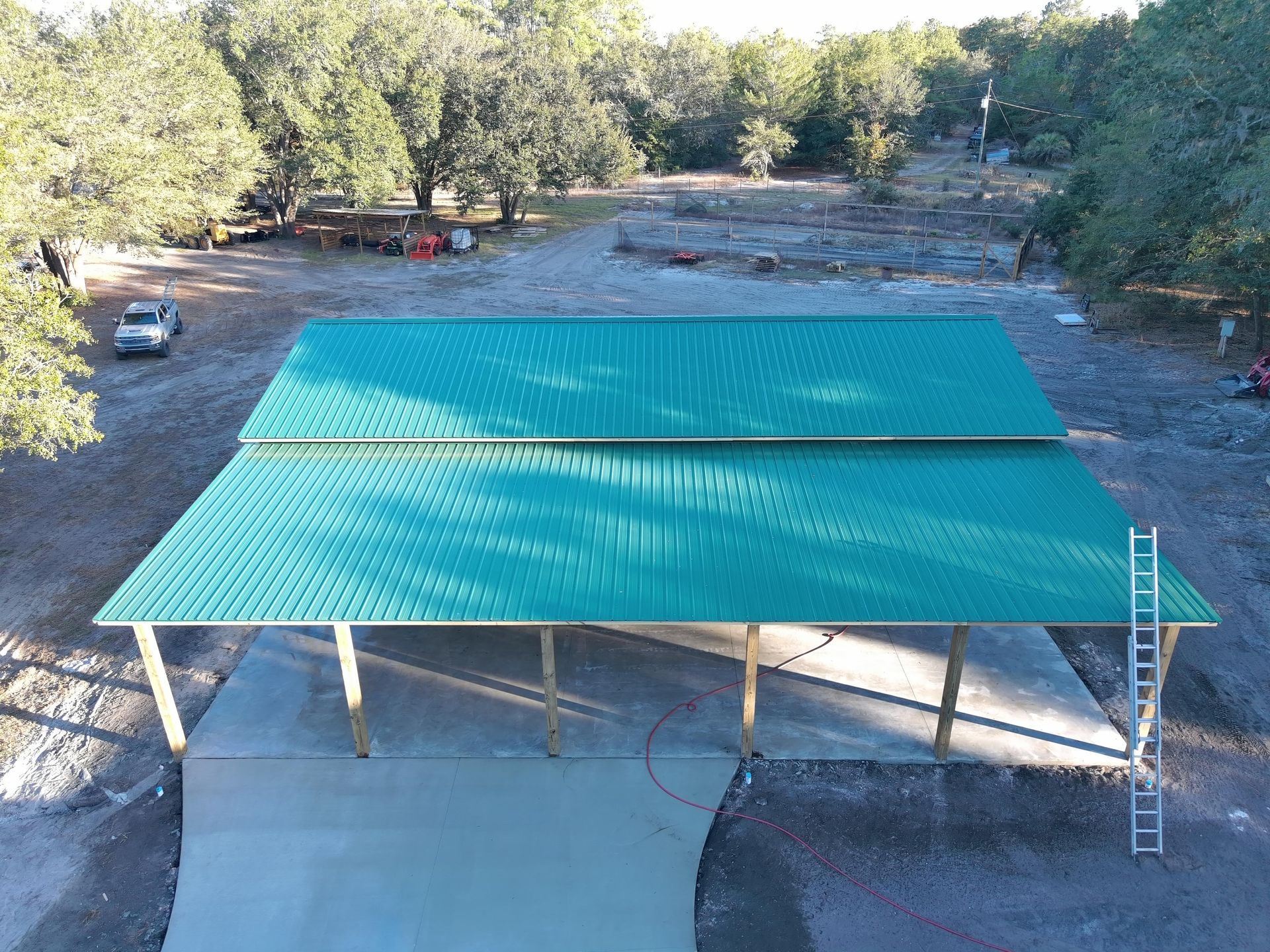 An elevated view of a large, green metal roof structure on a concrete slab in a wooded outdoor setting.