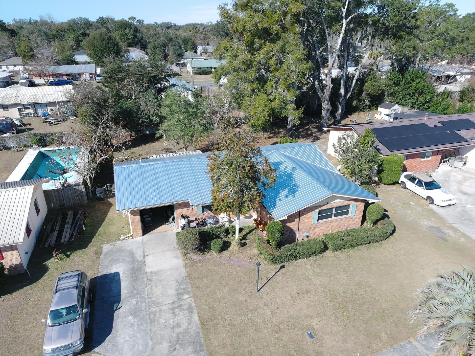 An aerial view of a brick house with a blue metal roof, a driveway with a car, and a swimming pool in a residential area.