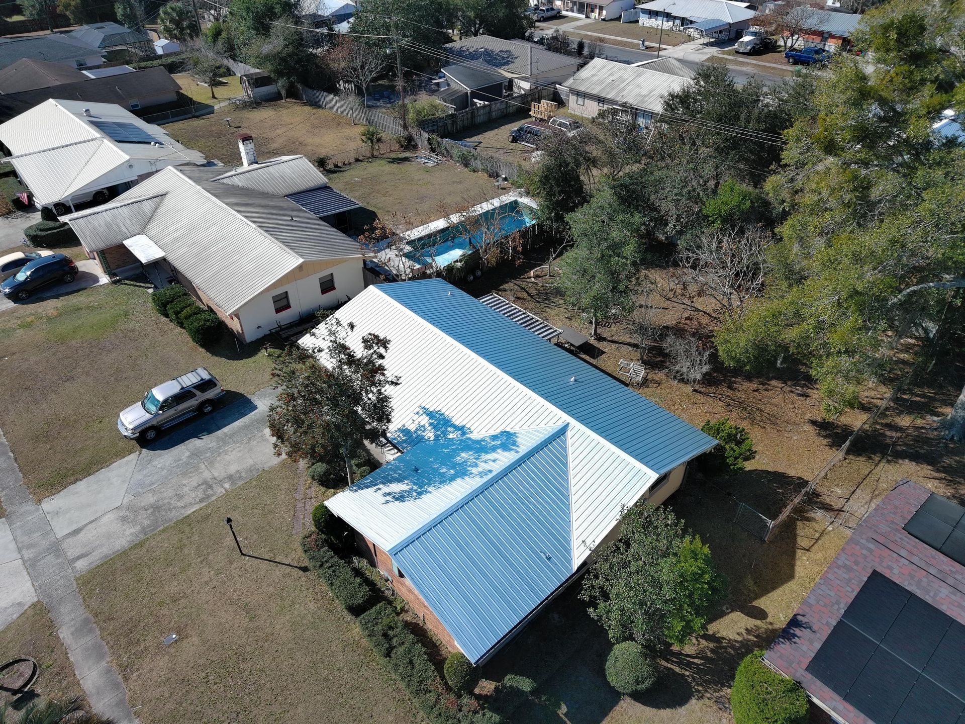 High-angle view of suburban houses, featuring a home with a blue and white metal roof, a driveway, and a backyard pool.